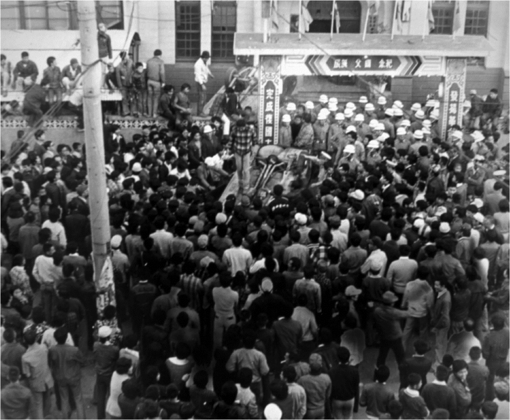 1977.11.19包圍中壢分局的群眾，其中一名站在翻倒的車子上演講。（771119N06／劉峰松提供） 